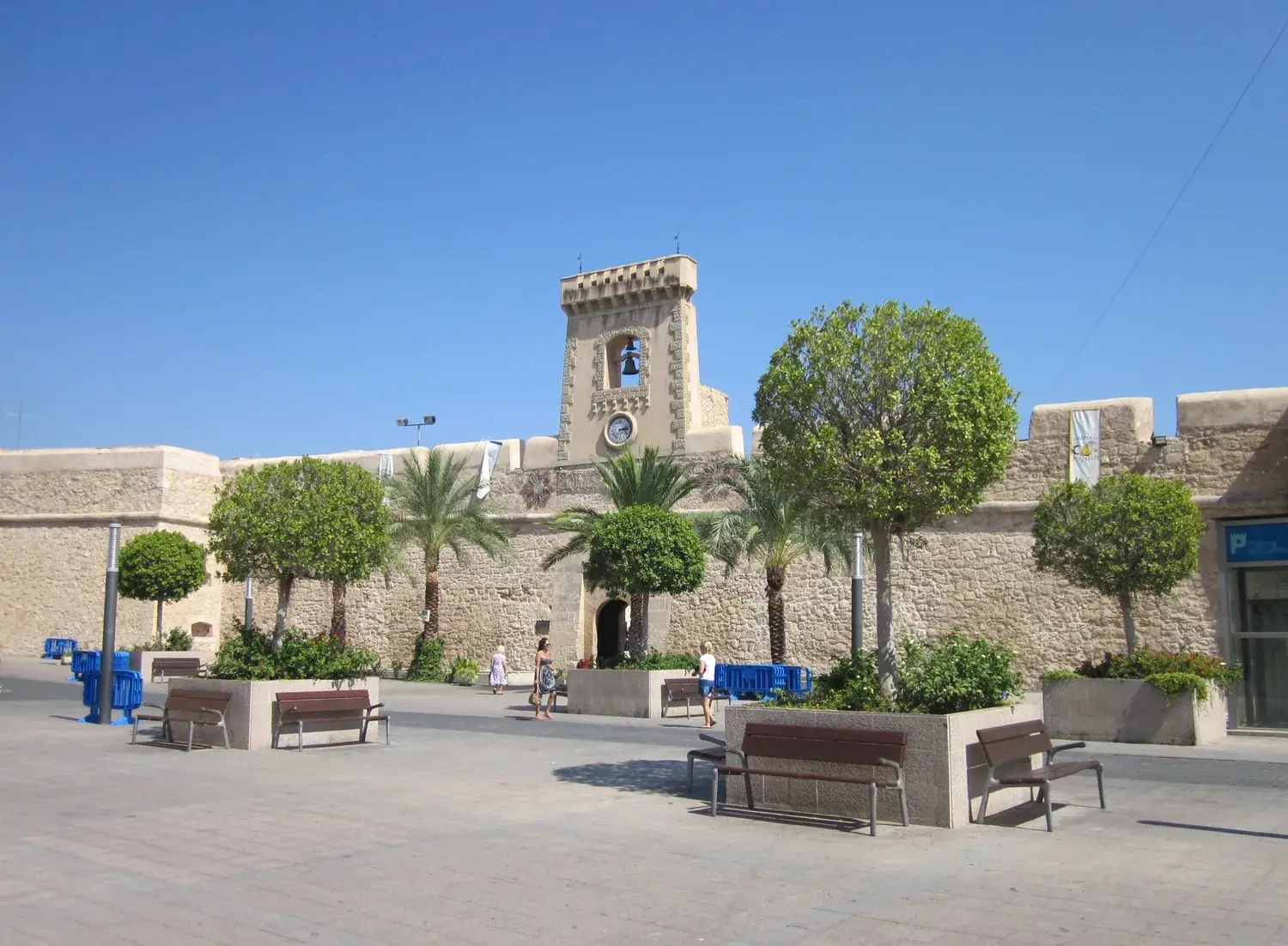 La forteresse de Santa Pola, ses murs de pierre et sa tour, sur une place plantée d’arbres