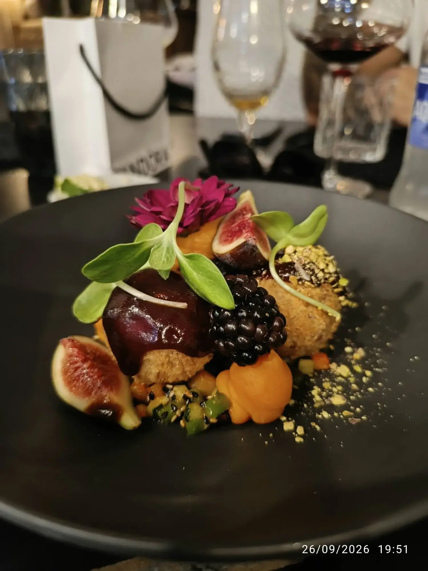 Croquetas de pato confit du Restaurante Luka dans une assiette noire, avec figues, mûre, purée orange, sauce sombre, pistaches concassées, jeunes pousses et une fleur violette