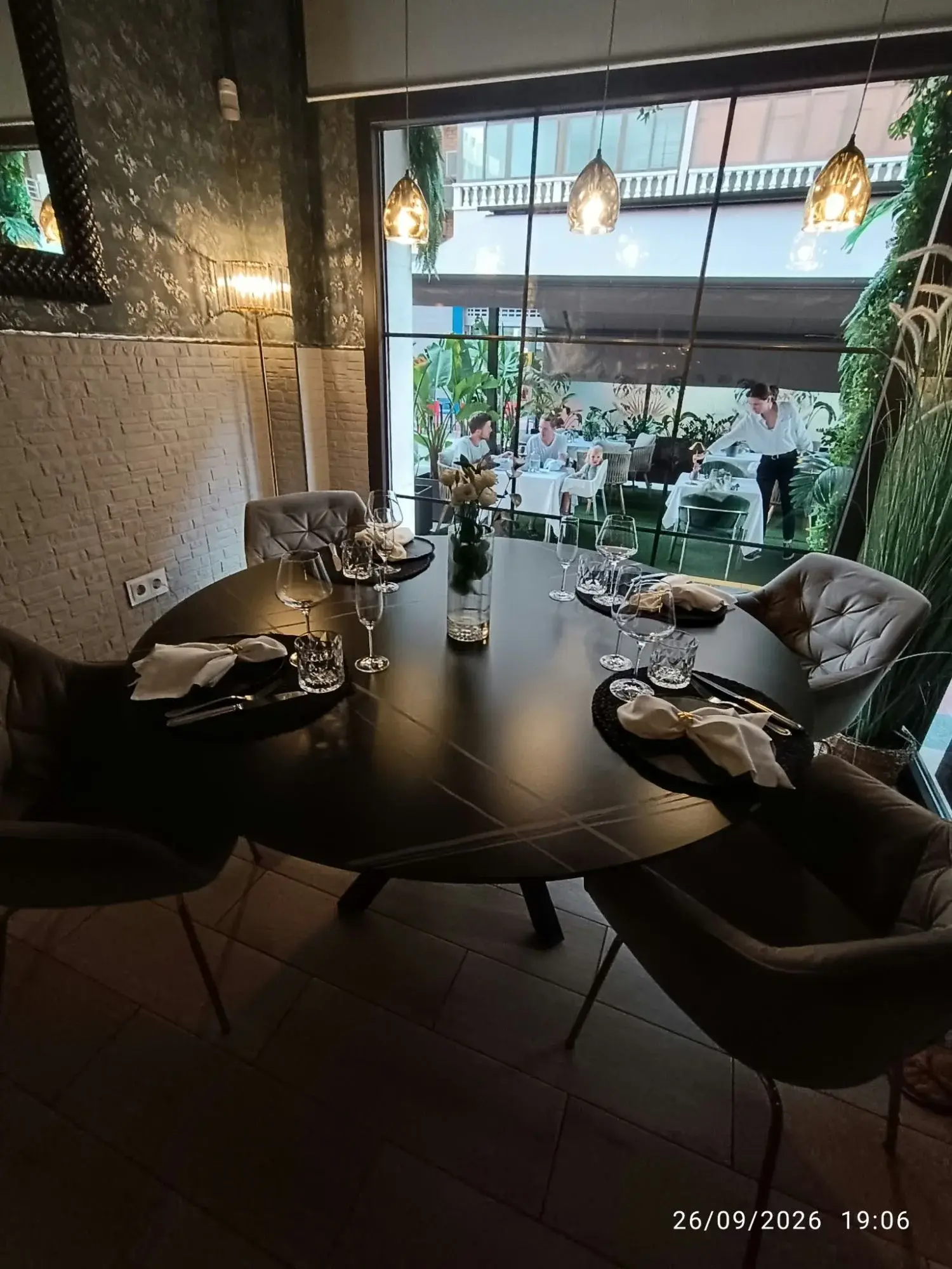 Table ronde dressée à l’intérieur du Restaurante Luka, avec verres à vin et serviettes nouées, et vue par la grande baie vitrée sur la terrasse végétalisée